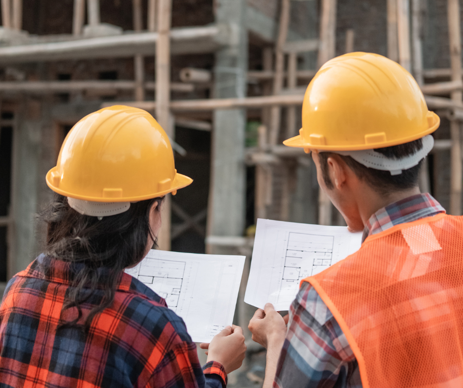Two contractors in flannel shirts and yellow hard hats face away from the camera as they look over blue prints of a building