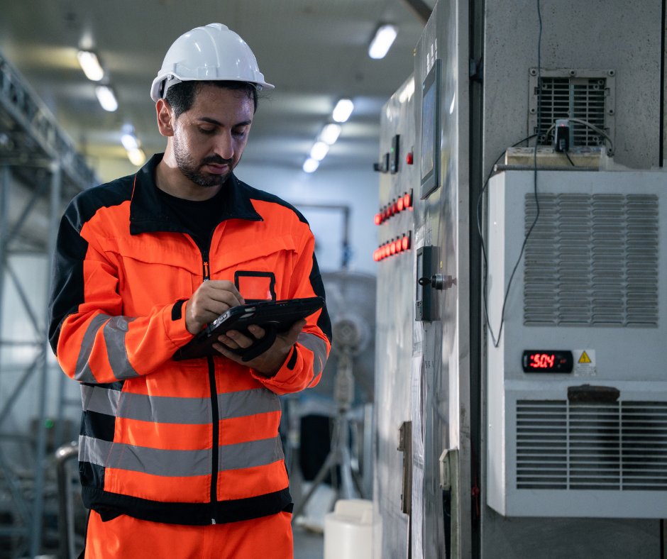 HVAC technician in bright orange uniform and white hard hat using tablet to check measurements of a system in a basement